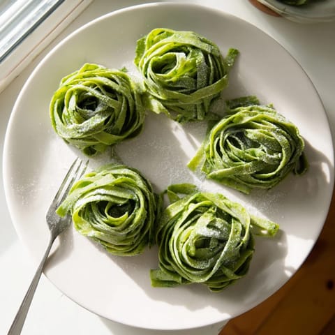 Fresh homemade spinach pasta dough being cut into delicate fettuccine ribbons on a wooden board.