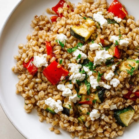 A close-up of golden, caramelized farro with roasted vegetables, including bell peppers and zucchini, steaming in a white ceramic bowl.